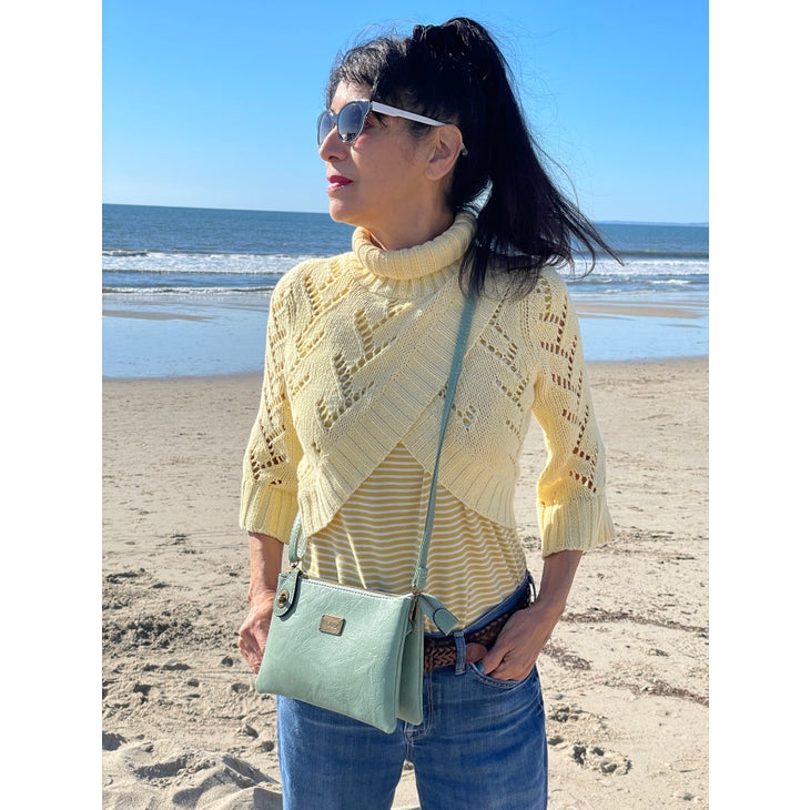 A woman stands on a beach wearing sunglasses, a yellow knit sweater over a striped shirt, and blue jeans. She carries the Tina Cell Bag in blush from Liz Soto Handbags, which can also be used as a wristlet. Her long dark hair is tied back in a ponytail, and the ocean stretches out behind her under the clear blue sky.
