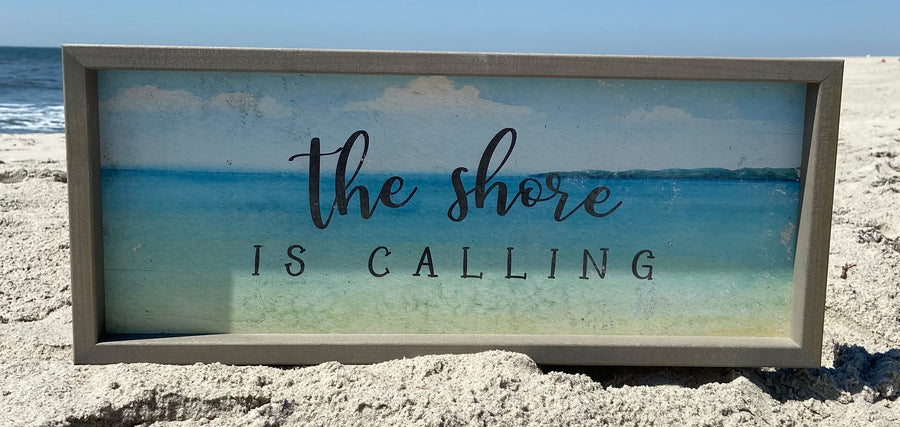 A framed sign, ready to hang, rests on the sandy beach with a serene ocean backdrop. Featuring an illustration of the sea and sky, it elegantly bears the words "The Shore Is Calling." The calm waves and clear blue sky enhance its charm, making this piece from SincereSurroundings the perfect size addition to any coastal decor.