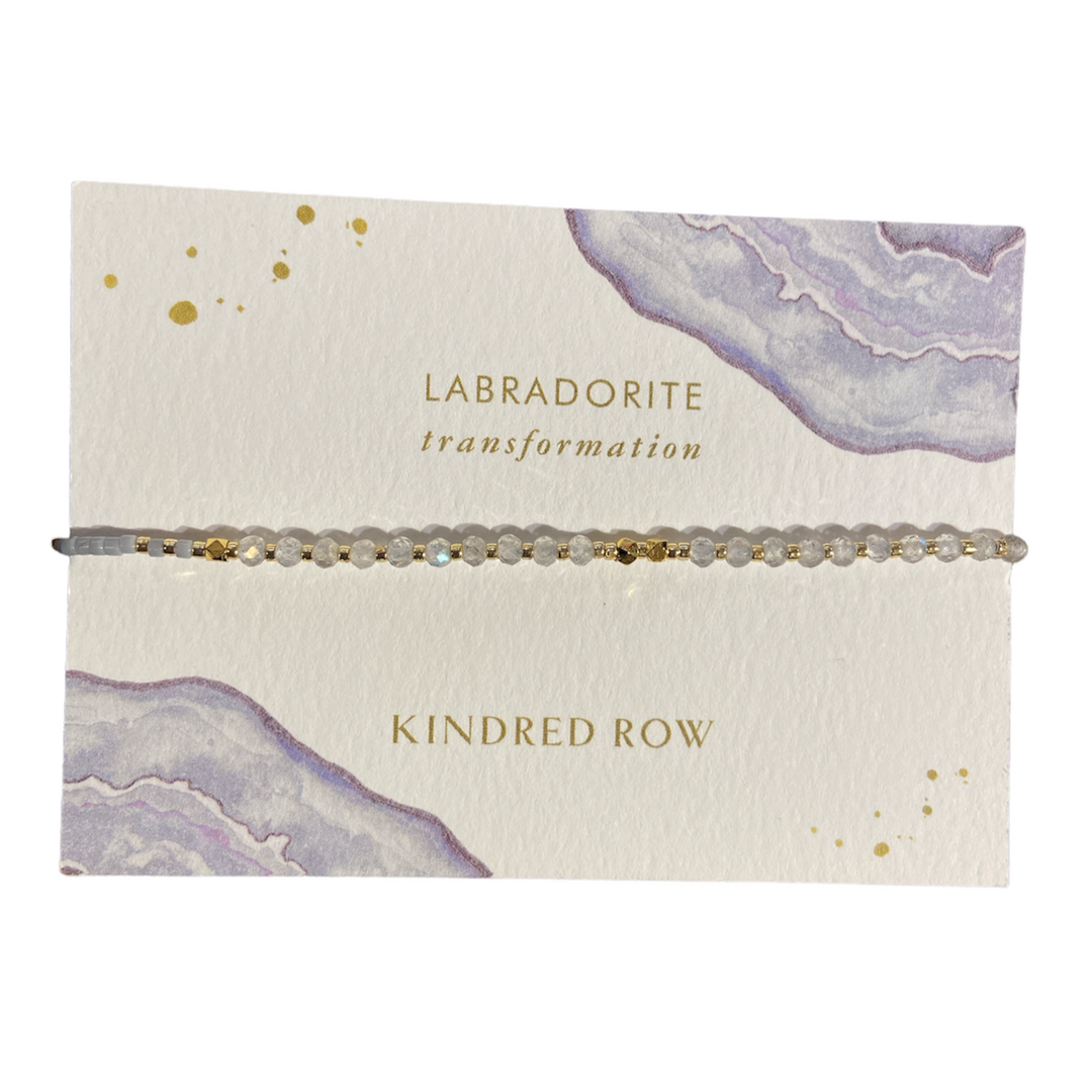 The Labradorite Healing Gemstone Stacking Bracelet by Kindred Row is a delicate, handmade piece displayed on a decorative card. The card showcases watercolor-like designs in shades of purple and gold, and includes the text "Labradorite Transformation" and "Kindred Row." This adjustable cotton cord bracelet is proudly made in the USA.