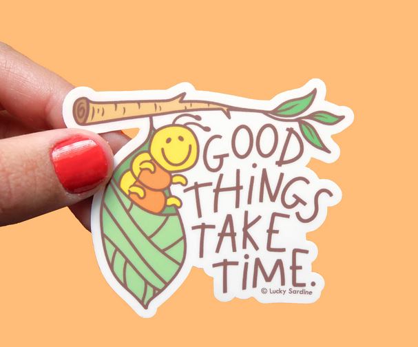 A hand with red nail polish holds a weatherproof vinyl sticker from Lucky Sardine against an orange background. The "Caterpillar Cocoon, Good Things Take Time" sticker features a smiling yellow cartoon snail on a branch with a leaf, alongside the phrase "Good things take time.