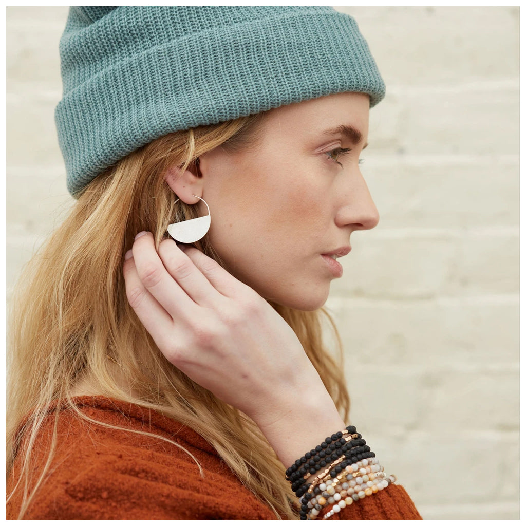 A person wearing a teal knitted hat, multiple beaded bracelets in various colors, and the Lunar Hoop Silver earrings by Scout Jewelry. They are dressed in a rust-colored sweater and have their hand up to their earring, looking to the side against a neutral brick wall background.