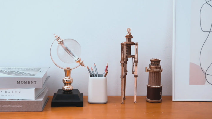 A neatly organized desk displays a stack of books, a glass sphere on a metallic stand, a cup holding colored pencils, two metallic robot models beside the DIY Wooden Puzzle: Monocular Telescope from Hands Craft, and part of a frame on a white wall. The overall aesthetic is minimalist with a touch of modern elegance.